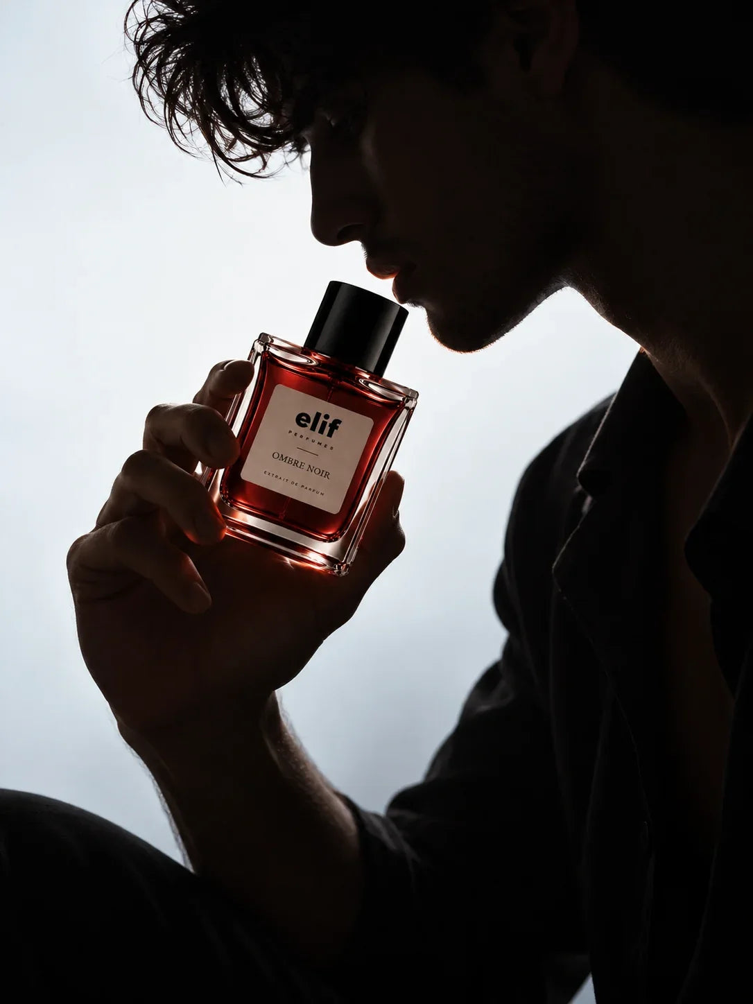 Elif Ombre Noir long-lasting perfume bottle held close to a man’s face as he smells the fragrance, shown in dramatic backlighting with a moody dark silhouette.