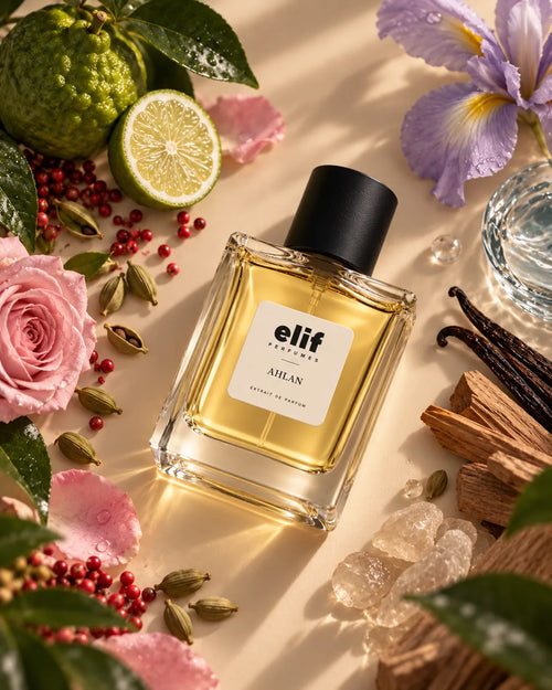 A bottle of Elif Perfumes 'Ahlan' fragrance with a black cap and clear glass, filled with golden-yellow liquid, styled on a beige surface with pink roses, a lime, green citrus, vanilla beans, and crystals.