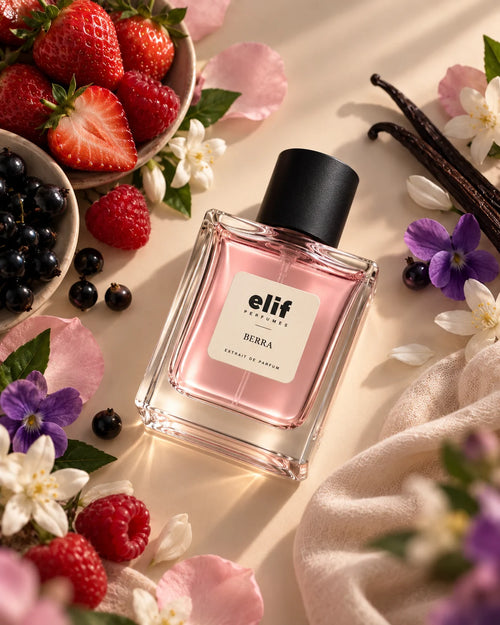 A bottle of Elif perfume, specifically the 'Berra' scent, is displayed on a warm beige surface surrounded by fresh strawberries, blueberries, vanilla beans, and delicate flowers. The perfume bottle is clear glass showing a soft pink liquid inside, topped with a matte black cap.