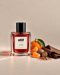 Elif Calor perfume bottle filled with deep red liquid and topped with a glossy black cap, displayed against a warm beige background. Fresh mandarin oranges and segments, cinnamon bark, dried spice, and green leaves are arranged beside the bottle, with warm directional lighting and subtle shadows. A long-lasting perfume designed for enduring wear.