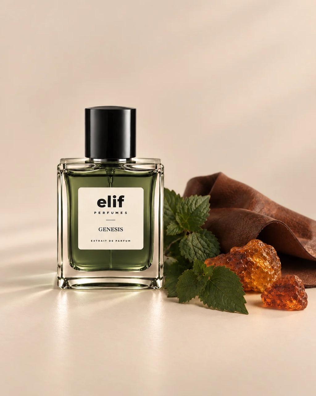 Elif Genesis perfume bottle with deep olive-green liquid and a black cap, displayed against a warm beige background. Fresh mint leaves, amber resin pieces, and a rich brown leather-like material are arranged beside the bottle, creating a warm, earthy composition. Soft lighting casts subtle shadows across the surface. A long-lasting fragrance with a fresh, green, warm, and resinous character.