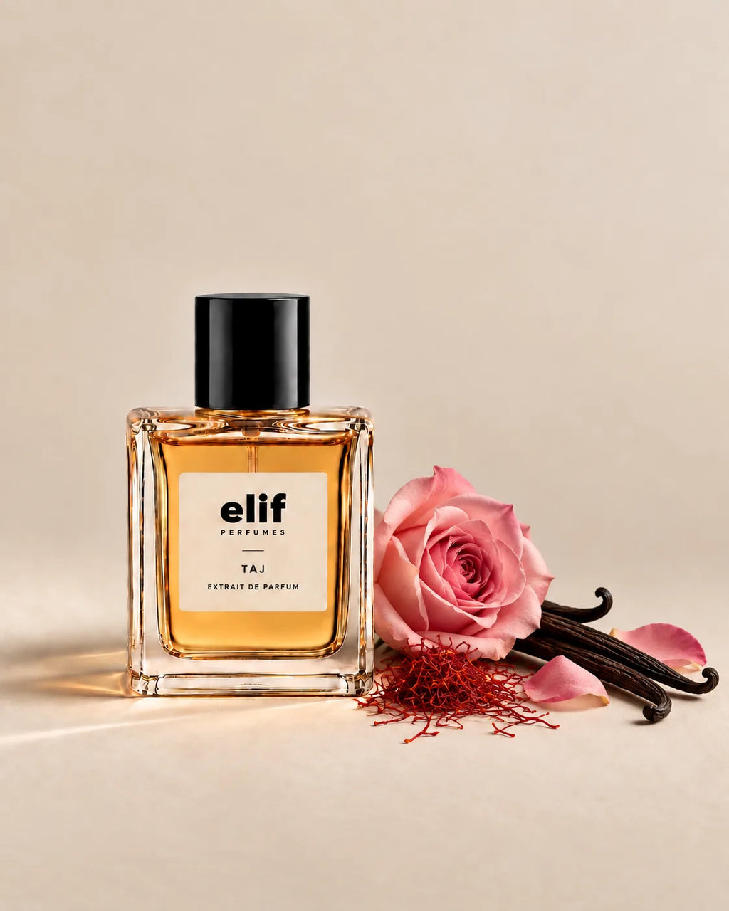 Elif TAJ perfume bottle filled with warm amber-gold liquid and topped with a glossy black cap, displayed against a warm beige background. A pink rose, vanilla pods, saffron threads, and scattered rose petals are arranged beside the bottle, with soft directional lighting and subtle shadows.