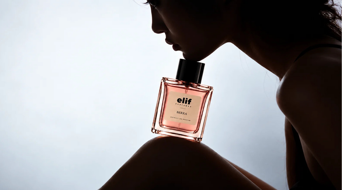 Elif Berra long-lasting perfume bottle held near a woman’s shoulder as she smells the fragrance, captured in dramatic backlighting with a soft silhouette and elegant pink-toned extrait de parfum bottle.